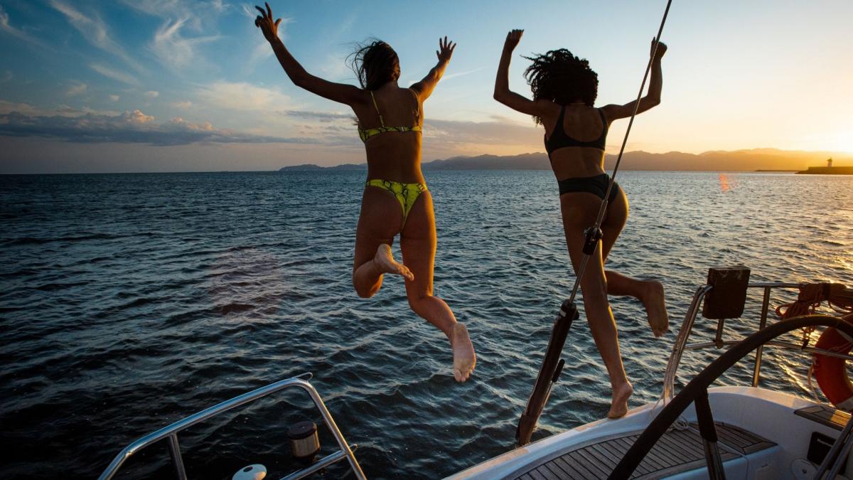 Zwei Frauen, die auf einem Boot Urlaub machen, springen bei Sonnenuntergang gemeinsam vom Deck ins Meer.